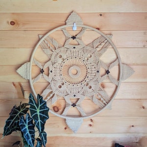 May include: A beige macrame wall hanging in a starburst design is mounted on a wooden wall. A potted plant with dark green leaves sits on a small black table. A black chair with a pink blanket is in the foreground.