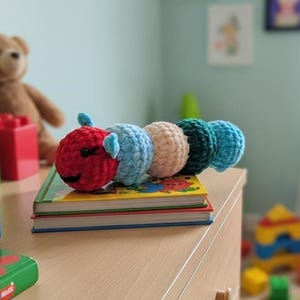 May include: A colorful, crocheted caterpillar toy with a red head, blue, tan, green, and blue body segments. The toy is resting on a stack of children's books on a wooden dresser. The background includes a teddy bear and other toys.