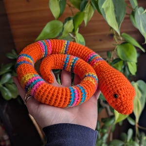 May include: A crocheted orange snake toy with colorful striped accents. The snake's body is a vibrant orange, with bands of blue, pink, yellow, and turquoise. The snake has a small black eye and is coiled.