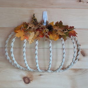 May include: A decorative pumpkin-shaped wall hanging made of twisted rope. The pumpkin is adorned with fall leaves in shades of orange, yellow, and red, along with pinecones and berries. The wall hanging is designed to be hung on a wall.