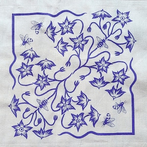 Op de afbeelding: Een vierkante, gebroken witte stof met een blauw bloemen- en bijenontwerp. Het ontwerp toont gestileerde bloemen en bijen binnen een golvende blauwe rand. De stof ligt op een houten oppervlak.