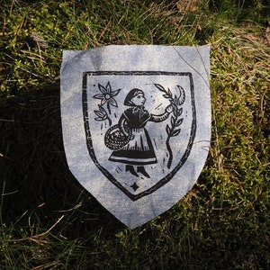 May include: A blue fabric shield with a black and white illustration of a person holding a basket and a flower. The shield is lying on a bed of green moss.