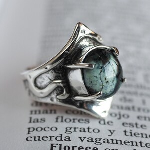 May include: A silver ring featuring a large, green gemstone set within an intricate, ornate design. The ring is displayed on a white, textured surface.