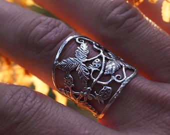 May include: A silver ring with a botanical design featuring leaves and berries. The ring is worn on a finger, with a backdrop of golden-hued foliage. The ring's intricate details are highlighted against the soft, natural lighting.