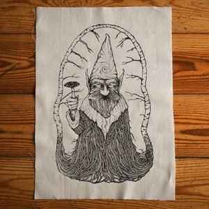 Puede incluir: Parche bordado en blanco y negro de una figura de gnomo con un sombrero puntiagudo, sosteniendo un hongo. La figura está rodeada por un diseño circular de ramas y enredaderas.