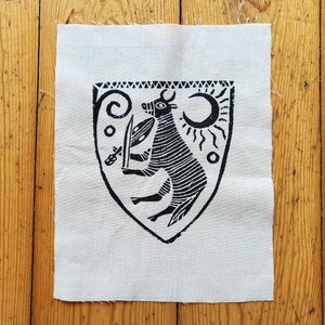May include: A black and white linocut print on a square piece of fabric. The print features a shield design with a bull-like figure holding a sword and shield, with a sun and crescent moon. The fabric is a light beige colour.