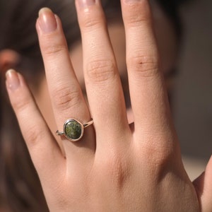 May include: A silver ring featuring a green oval-shaped gemstone. The ring is worn on a person's finger.