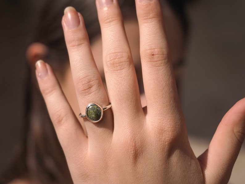 May include: A silver ring featuring a green oval-shaped gemstone. The ring is worn on a person's finger.
