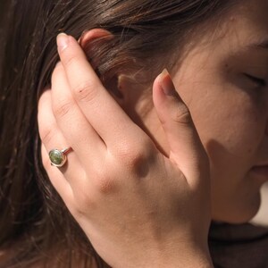 May include: A silver ring with a green oval gemstone. The ring is on a person's finger.