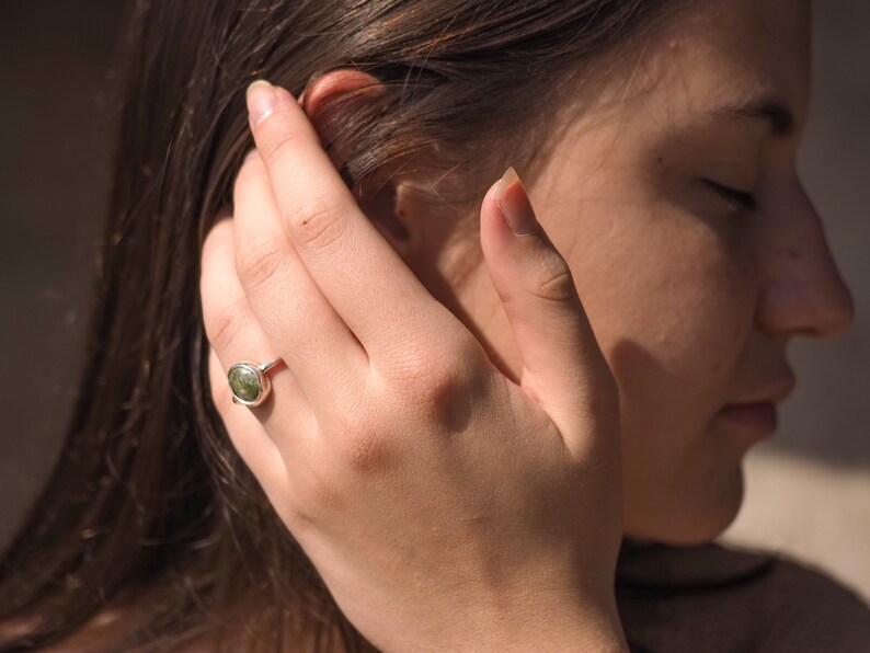 May include: A silver ring with a green oval gemstone. The ring is on a person's finger.