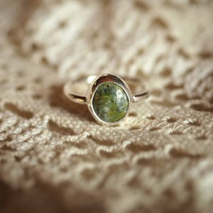 May include: A silver ring with a green oval-shaped gemstone set in the centre. The ring is on a white crocheted surface.