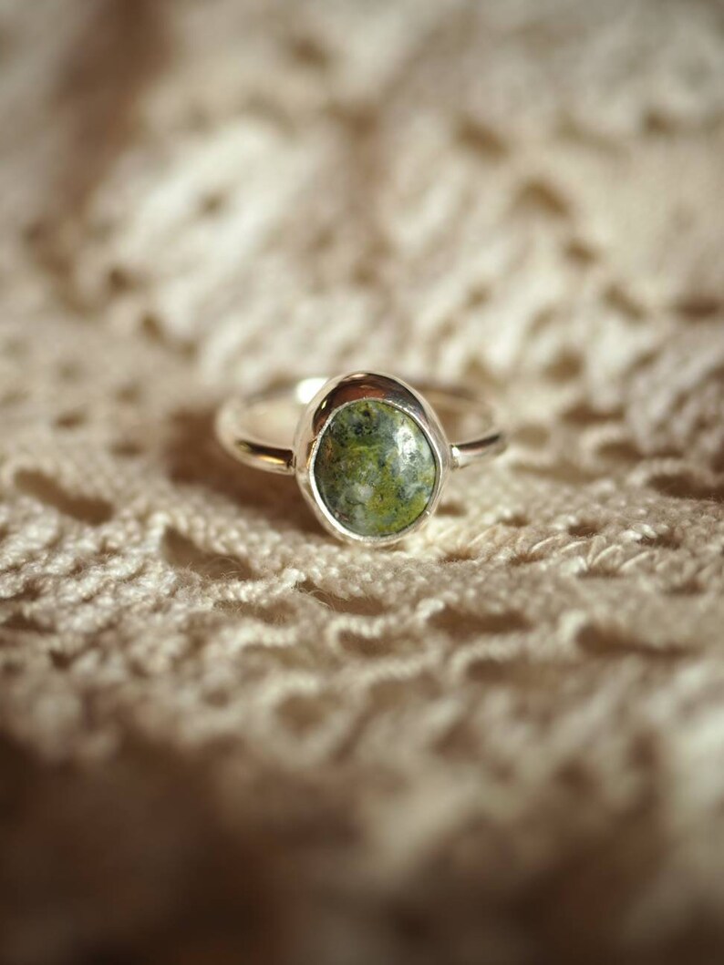 May include: A silver ring with a green oval-shaped gemstone set in the centre. The ring is on a white crocheted surface.
