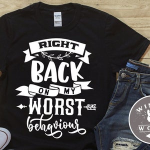 Può includere: Maglietta nera con testo bianco che recita "Right Back On My Worst Behaviour". La maglietta è stesa su una superficie di legno con un paio di jeans blu e una scarpa da ginnastica bianca.