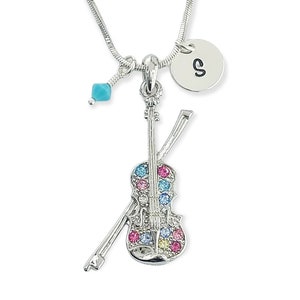 May include: Silver-toned necklace featuring a violin pendant adorned with colorful gemstones. The pendant includes a bow and is accented with a turquoise bead and a disc charm with the letter "S". The chain is a delicate snake chain.