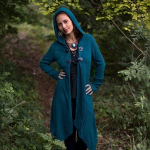 Peut inclure: Un cardigan à capuche bleu sarcelle avec une longue ligne d'ourlet asymétrique et une rangée de boutons noirs sur le devant. Le cardigan est fait d'un tissu doux et tricoté et a une coupe décontractée.