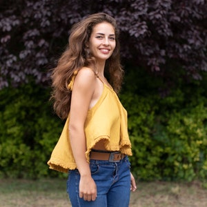 May include: A woman with long brown hair is wearing a yellow sleeveless top with a fringe trim and a brown belt. She is smiling and looking at the camera.