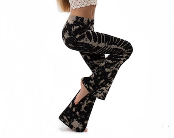 May include: A woman wearing a white crochet top and black and white tie-dye bell bottom pants. She is standing on one leg with her arms raised in a yoga pose.