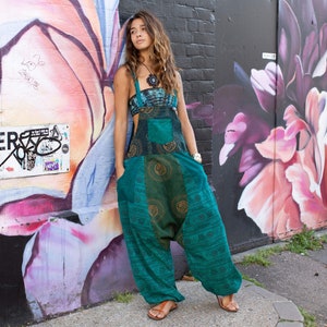 Może przedstawiać: Kobieta w turkusowym kombinezonie z wzorzystym wzorem. Kombinezon ma szerokie nogawki i luźny krój. Kobieta stoi przed ceglaną ścianą z graffiti.