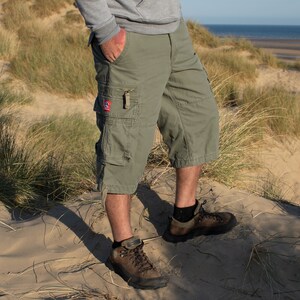 May include: A person wearing green cargo shorts and brown hiking boots standing on a sandy beach with tall grass.