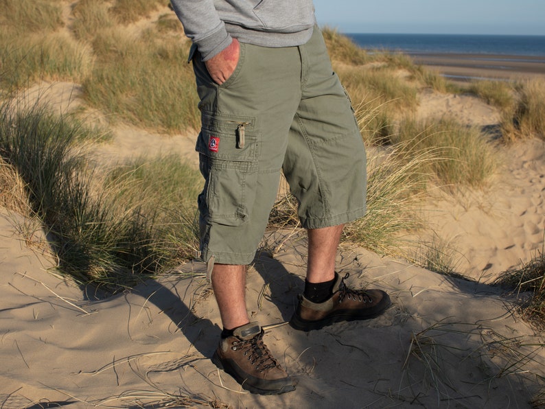 May include: A person wearing green cargo shorts and brown hiking boots standing on a sandy beach with tall grass.
