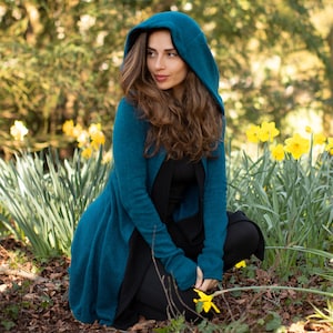 Puede incluir: Una mujer con un cárdigan azul turquesa con capucha se sienta en un campo de narcisos amarillos. El cárdigan tiene un diseño largo y fluido y está hecho de un tejido suave y de punto.