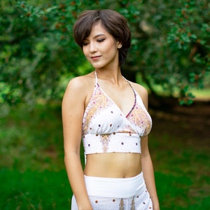 Puede incluir: Un top blanco de tirantes con estampado de plumas de pavo real de colores. El top tiene un escote en V y un cierre de nudo en la espalda.