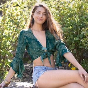 May include: A teal green paisley print crop top with bell sleeves and a tie front. The top is paired with denim shorts. The model is wearing gold bracelets and rings. The background features green foliage.