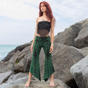 May include: A woman wearing a black tube top and green patterned pants stands on rocks near the ocean. The pants have a leaf-like design in black on a green background. The top is a sheer black material.