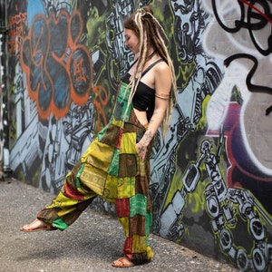 Może przedstawiać: Kobieta w kolorowym kombinezonie w stylu patchwork, czarnym topie na ramiączkach i długimi blond dredami. Stoi przed ścianą z graffiti.