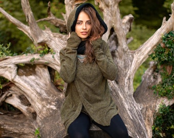 May include: An olive green hooded sweater with a black trim and a decorative emblem. The sweater has long sleeves and a wrap-style front. The person is wearing a black scarf and black pants. The background features a tree trunk and foliage.