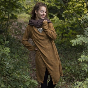 May include: A woman wearing a long, brown, knitted cardigan with a brown scarf around her neck. She is standing in a forest setting.