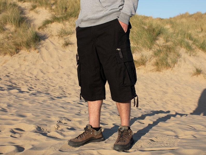 May include: A man wearing black cargo shorts with multiple pockets and brown hiking boots. He is standing on a sandy beach.