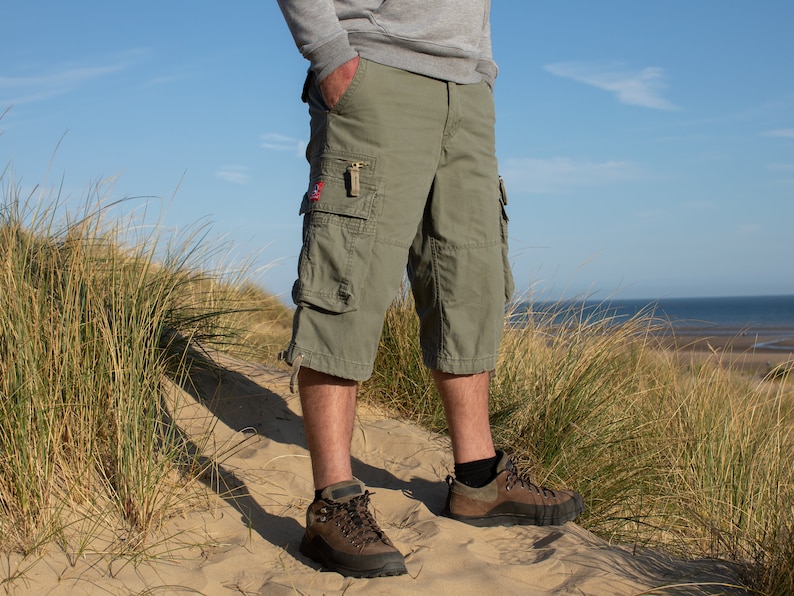 May include: A man wearing green cargo shorts and brown hiking boots stands on a sand dune. The shorts have a large cargo pocket on the left leg and a smaller pocket on the right leg.