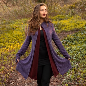 May include: A long, purple cardigan with a burgundy lining. The cardigan has a hood and long sleeves with thumbholes. The model is wearing the cardigan with a black top and black leggings.