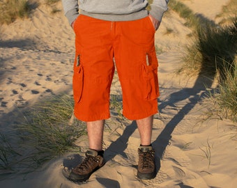May include: A pair of orange cargo shorts with multiple pockets. The shorts are worn by a person standing on a sandy beach.