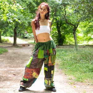 Peut inclure: Pantalon vert à patchwork avec taille élastique. Le pantalon présente divers patchs de tissu dans les tons verts, marron et orange. Un haut tube blanc est également visible. La personne se tient sur un chemin de terre dans un parc.