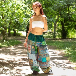 May include: A person wearing a white tube top and patchwork pants. The pants are a mix of green, blue, and tan fabrics with various patterns. The pants have a drawstring waist and a loose, comfortable fit. The background is a park setting.
