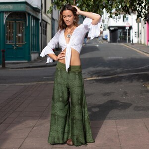 May include: A woman wearing a white crop top with a tie front and green printed wide-leg pants. She is standing on a sidewalk in front of a building.