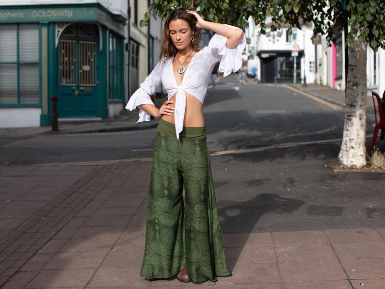 May include: A woman wearing a white crop top with a tie front and green printed wide-leg pants. She is standing on a sidewalk in front of a building.
