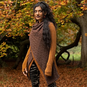 May include: A long-sleeved sweater in brown and orange with a cowl neck and a draped front. The sweater is being worn by a person with long dark hair standing in a woodland setting with autumn leaves.