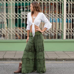 May include: A woman wearing a white crop top with a tie front and green printed wide-leg pants. She is standing in front of a green wall with a metal gate behind her.