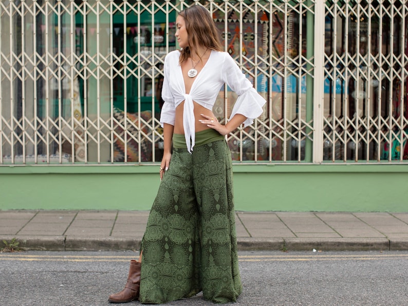 May include: A woman wearing a white crop top with a tie front and green printed wide-leg pants. She is standing in front of a green wall with a metal gate behind her.