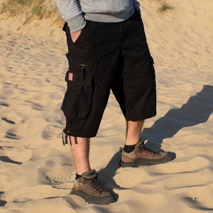 May include: Black cargo shorts with a red patch on the left leg. The shorts are worn by a person standing on a sandy beach.