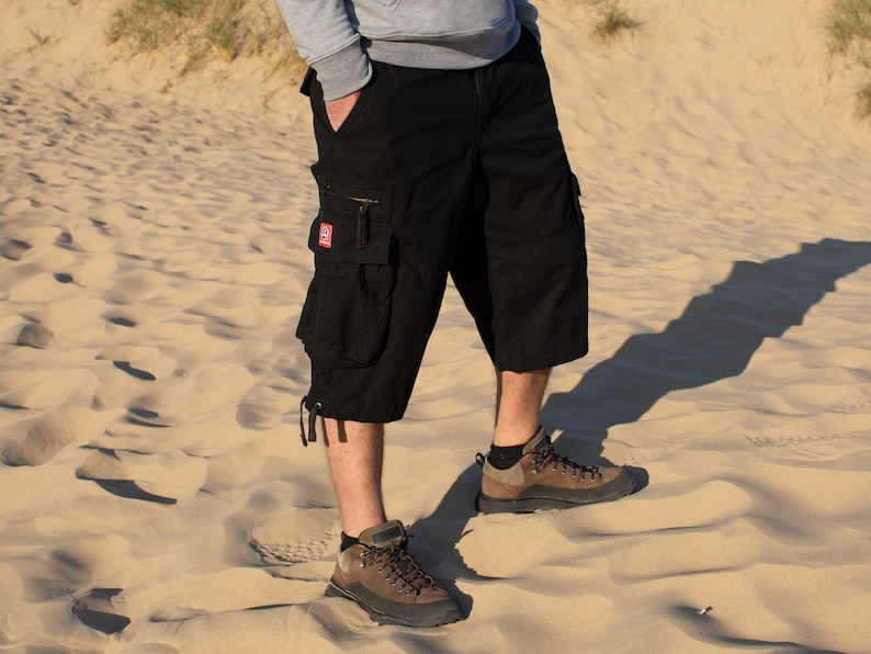 May include: Black cargo shorts with a red patch on the left leg. The shorts are worn by a person standing on a sandy beach.