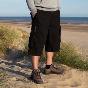 May include: A person wearing black cargo shorts with multiple pockets and brown hiking boots. The person is standing on a sandy beach with tall grass in the foreground.