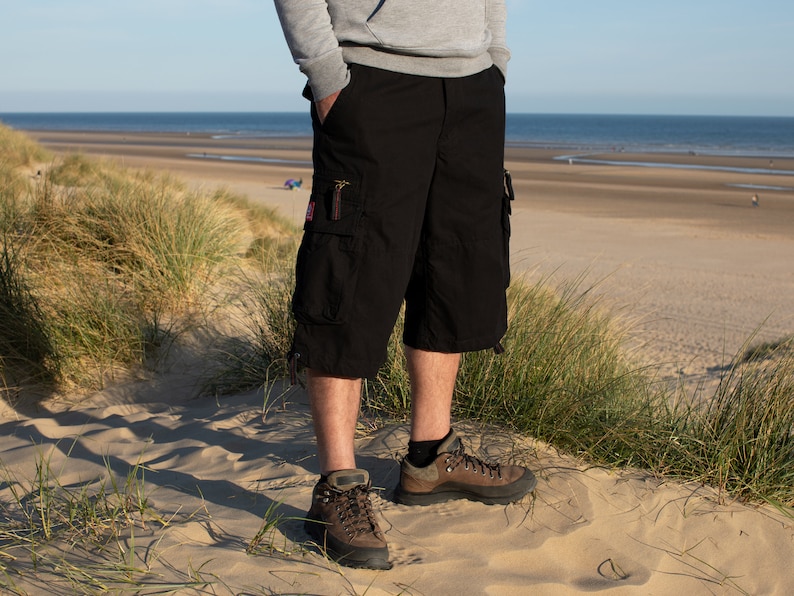 May include: A person wearing black cargo shorts with multiple pockets and brown hiking boots. The person is standing on a sandy beach with tall grass in the foreground.