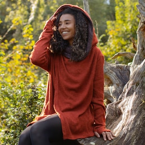 Puede incluir: Una mujer con un suéter con capucha de color óxido con mangas largas. Está sentada en un tronco de árbol grande y desgastado en un entorno boscoso.