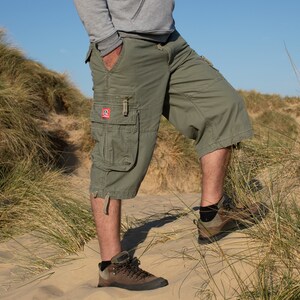May include: A person wearing green cargo shorts with a patch on the left leg. The person is standing on a sandy beach with tall grass in the background.