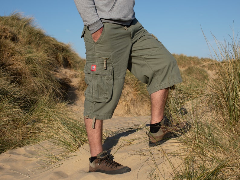 May include: A person wearing green cargo shorts with a patch on the left leg. The person is standing on a sandy beach with tall grass in the background.