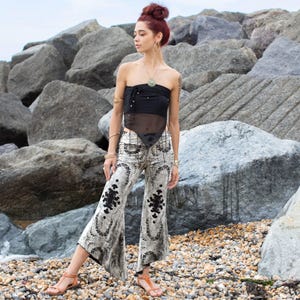 Puede incluir: Un top negro sin tirantes con un panel transparente y pantalones acampanados estampados. Los pantalones son principalmente blancos con un diseño floral negro. La modelo lleva joyas doradas y sandalias naranjas. El fondo presenta grandes rocas grises y una playa.